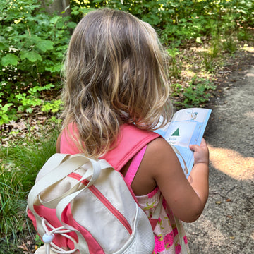 Child with a backpack and pink dress standing outdoors on a path.