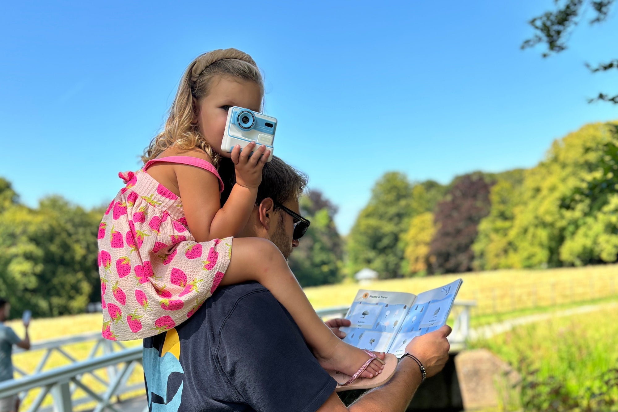 Man holding a child on his shoulders by a lake, both looking at a map.