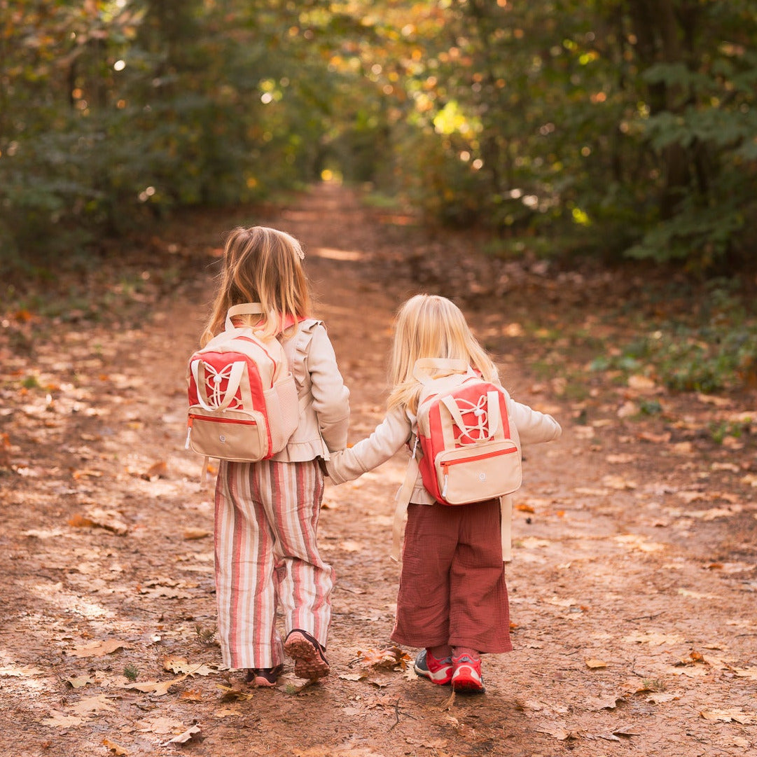 Two children walking down a leaf-covered path in a forest with backpacks.