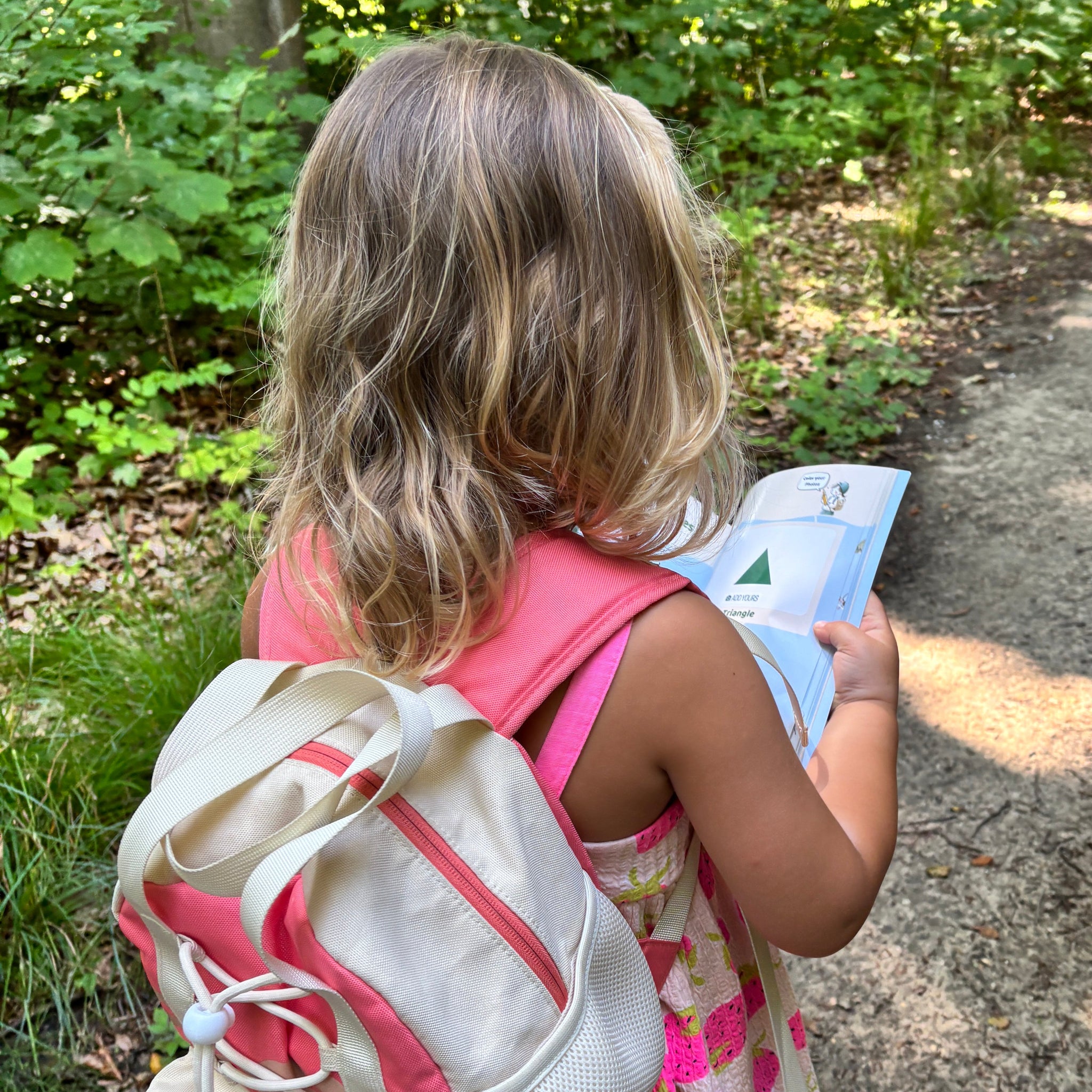 Child with a backpack and pink dress standing outdoors on a path.