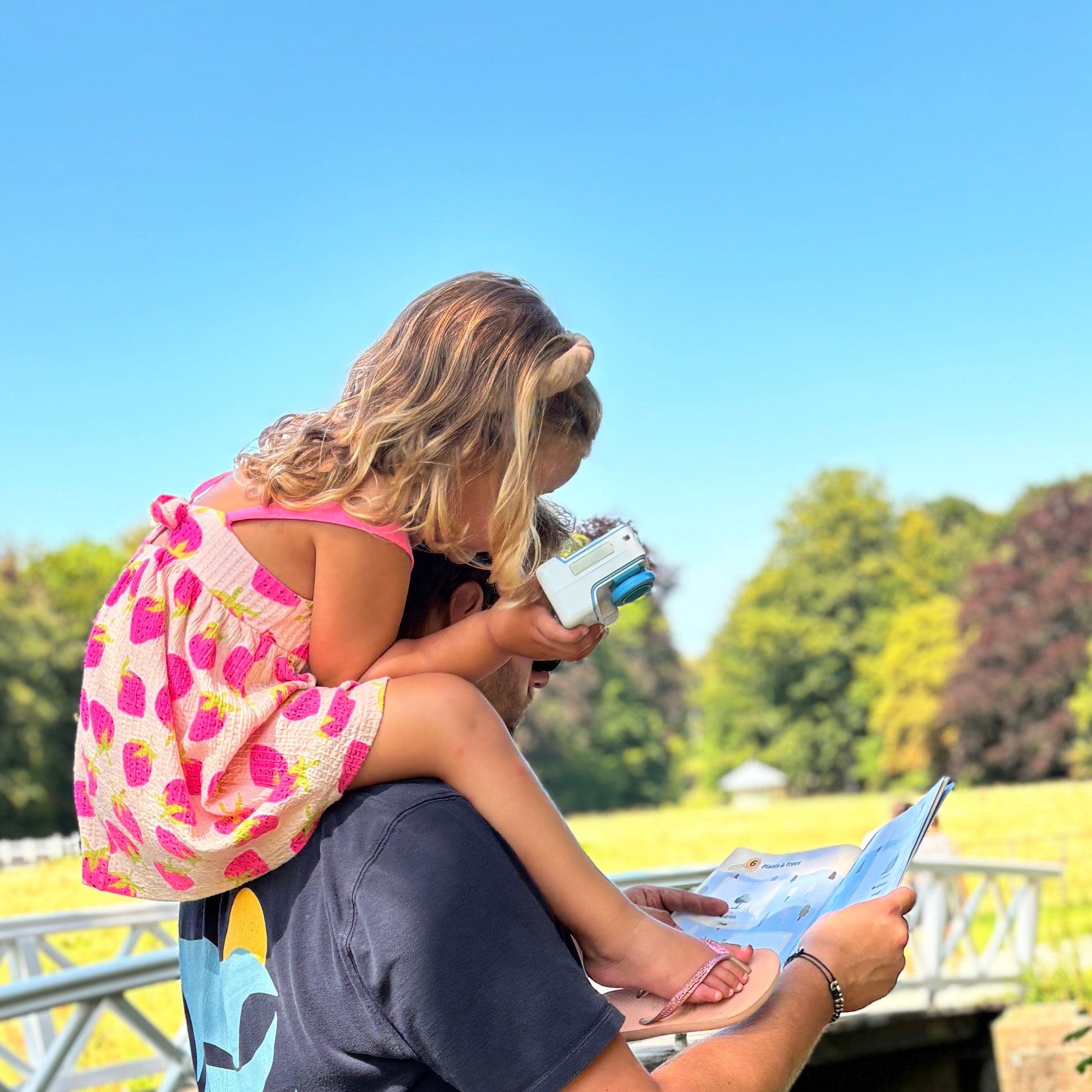Man with child on shoulders by a canal, looking at an adventure album.