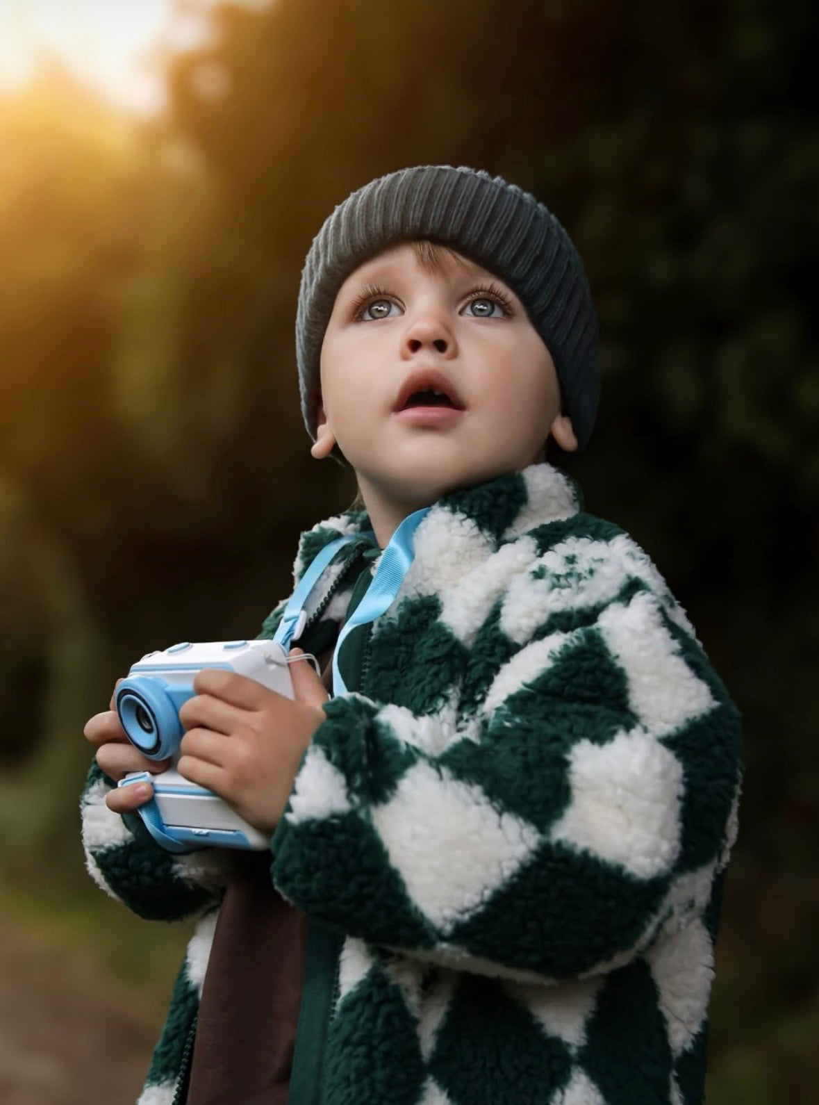 Child wearing a checkered coat and knit hat, holding a toy camera outdoors from Trafello.